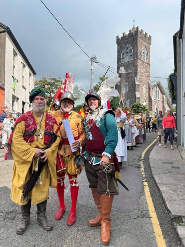 Imagen de personas vestidas con trajes medievales en una calle con edificios históricos y un campanario.