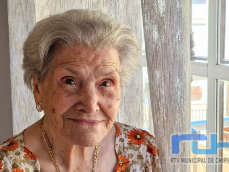 Una anciana con cabello gris y una sonrisa amable, vestida con un top floral. Detrás de ella se ve una ventana con barrotes y una puerta de madera.