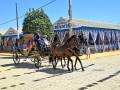 Imagen de un carruaje con dos caballos en el centro, conduciendo a una persona vestida de azul y otro de rojo. El carruaje está en frente de una tienda con la palabra "Bendita" y otra con "Gloria". La escena se desarrolla en un día soleado, con un cielo azul claro y una línea de árboles en el fondo.