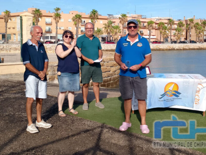 Grupo de personas posando frente a un lago con edificios y palmeras en el fondo. En primer plano, una mesa con un cartel de la ciudad de Chipiona. El cielo es claro y el ambiente parece ser de una mañana soleada en un entorno urbano con elementos naturales.