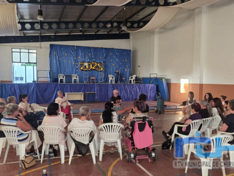 Una sala de conferencias con asistentes sentados en sillas blancas, un escenario con una bandera azul y estrellas doradas, y una pantalla de proyección en la pared.