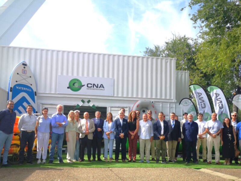 Una imagen de un grupo de personas posando frente a una instalación industrial con el logo CNA. El equipo está formado por hombres y mujeres vestidos de manera formal, posando en una zona verde con árboles al fondo. En el lado izquierdo, hay banderas de CNA y un paraguas con la marca. La construcción tiene una fachada blanca con detalles grises y azules, y el cielo está despejado.