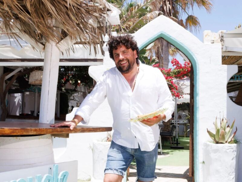 Un hombre con una camisa blanca y pantalones cortos está sonriendo mientras sostiene un bollo de pan en una barra de madera. Detrás de él, se puede ver un toldo de paja y una planta con flores rojas. El fondo muestra un ambiente tropical con palmeras y una estructura blanca de madera.