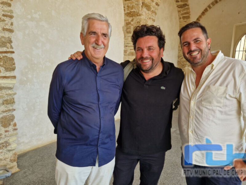 Tres hombres sonrientes posan juntos en una entrada de piedra. La imagen está acompañada por el logo de la RTV Municipal de Chipiona en la esquina inferior derecha.