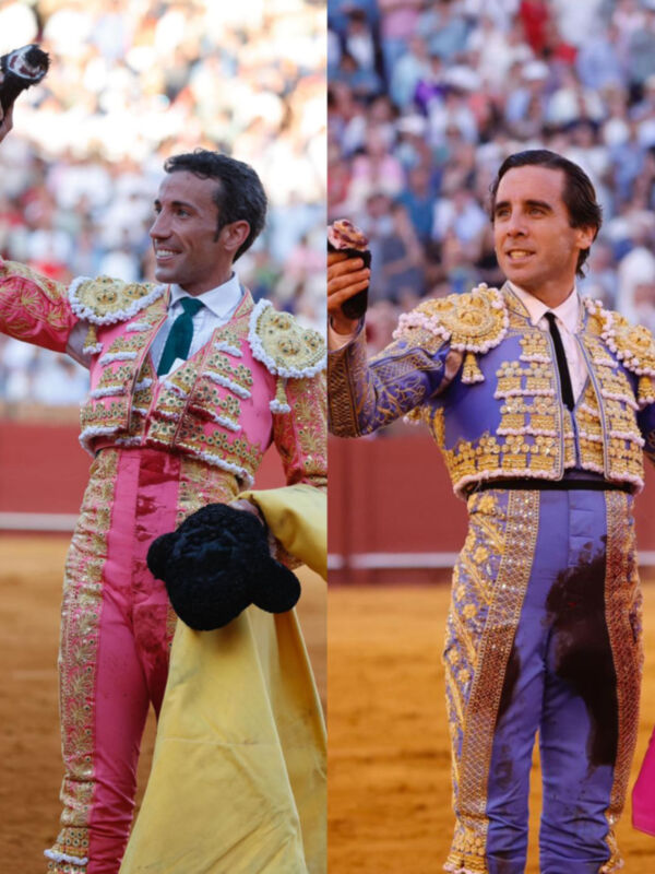 Dos toreros en trajes tradicionales de color rosa y azul, respetivamente. Ambos sostienen capotes amarillos en sus manos. El fondo muestra una multitud en un estadio lleno, probablemente durante una corrida de toros.