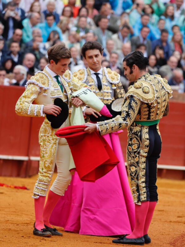 Un torero en traje de lujo dorado y blanco se prepara para una corrida, mientras un joven ayudante lo asiste. El fondo muestra a un público en el estadio.