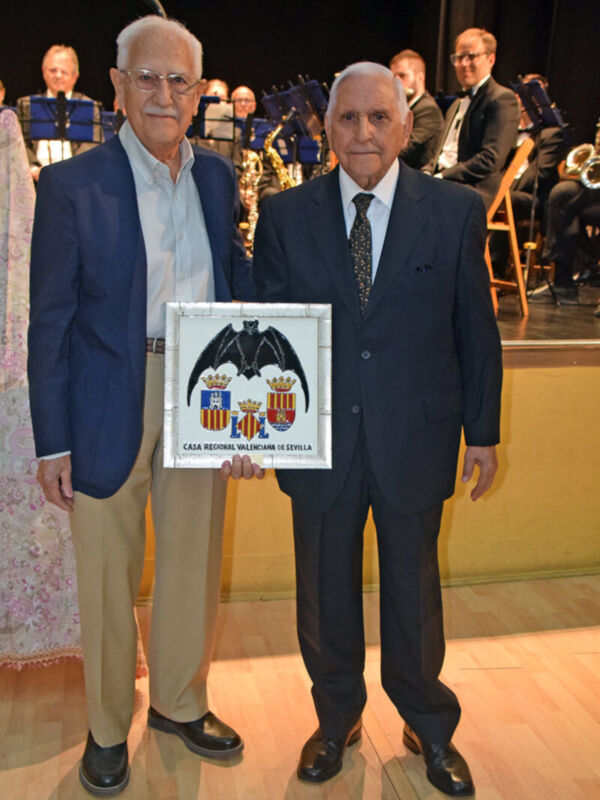 Dos hombres mayores posan con un reconocimiento de la Caja Regional Mallorquina. El fondo muestra a una orquesta musical en un escenario iluminado.