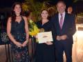 Una mujer en un vestido largo y una mujer en un vestido negro sostienen un diploma mientras un hombre en traje y corbata lo mira. El fondo muestra una terraza con palmeras y luces.