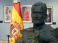 Busto de un hombre vestido con armadura, frente a una bandera española. En el fondo, dos retratos de personas en rojo y azul.