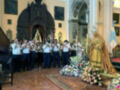 Imagen de una iglesia con un coro de músicos tocando instrumentos tradicionales. La arquitectura es elegante con detalles dorados y una estatua de la Virgen en el centro. El suelo es de mármol y hay flores blancas y rosadas alrededor.