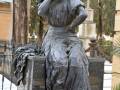 Estatua de bronce de una mujer sentada en un monumento, con cabello largo y vestida con un vestido antiguo. La escultura se encuentra en un cementerio rodeada de árboles y otros monumentos.