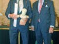 Dos hombres en trajes posan con un busto de bronce y un diploma, probablemente en una ceremonia de premiación.