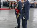 Imagen de oficiales militares en uniforme, posando formalmente. En el fondo, se observan otros oficiales y banderas en un hangar o edificio industrial.