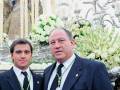 Dos hombres en trajes formales posan frente a una estatua religiosa y un gran arreglo floral blanco.